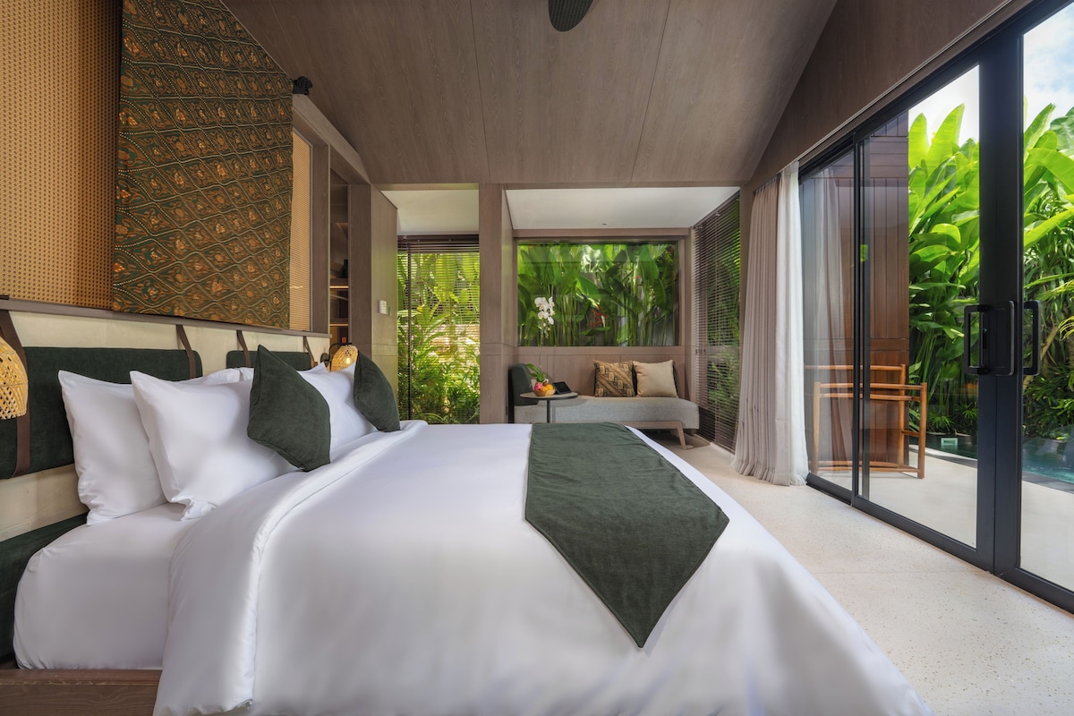 Un dormitorio moderno con una cama grande, un pequeño espacio de descanso y un ventanal corredero a un jardín exuberante.
