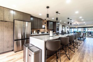 A modern kitchen with a stainless steel refrigerator, wooden cabinets, a central island with bar stools, and pendant lighting. - Beachfront Bliss Panoramic Ocean Views Sleeps 33 (Corpus Christi)