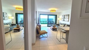 Una sala moderna con sofá, mesa de centro y sillón. Hay una zona de comedor con mesa y sillas. La habitación tiene un ventanal con vistas al mar.