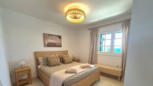 Un dormitorio con cama, mesita de noche, lámpara, ventana con cortinas y una obra de arte colgada en la pared.