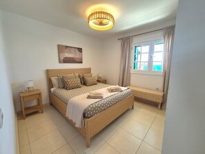 Ein Schlafzimmer mit Bett, Nachttisch, Lampe, Fenster mit Vorhängen und einem Wandbild.