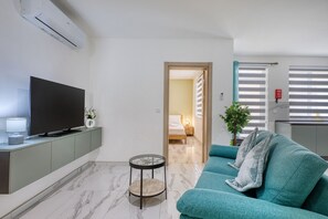 Ein modernes Wohnzimmer mit einem Flachbildfernseher, einem türkisfarbenen Sofa, einem kleinen runden Tisch und einem Flur, der zum Schlafzimmer führt.