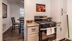 A kitchen with a stove, oven, and cabinets, a dining area with chairs, and a painting on the wall. - Blue Wave Cottage (Corpus Christi)