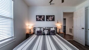 A bedroom with a large bed, two bedside lamps, a window with blinds, and two wall-mounted artworks. - Blue Wave Cottage (Corpus Christi)