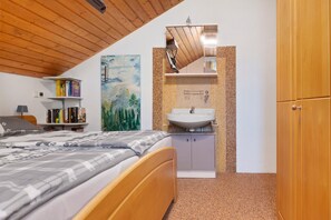 Un dormitorio con cama, mesita de noche, estantería, un cuadro, baño con lavabo y armario de madera.