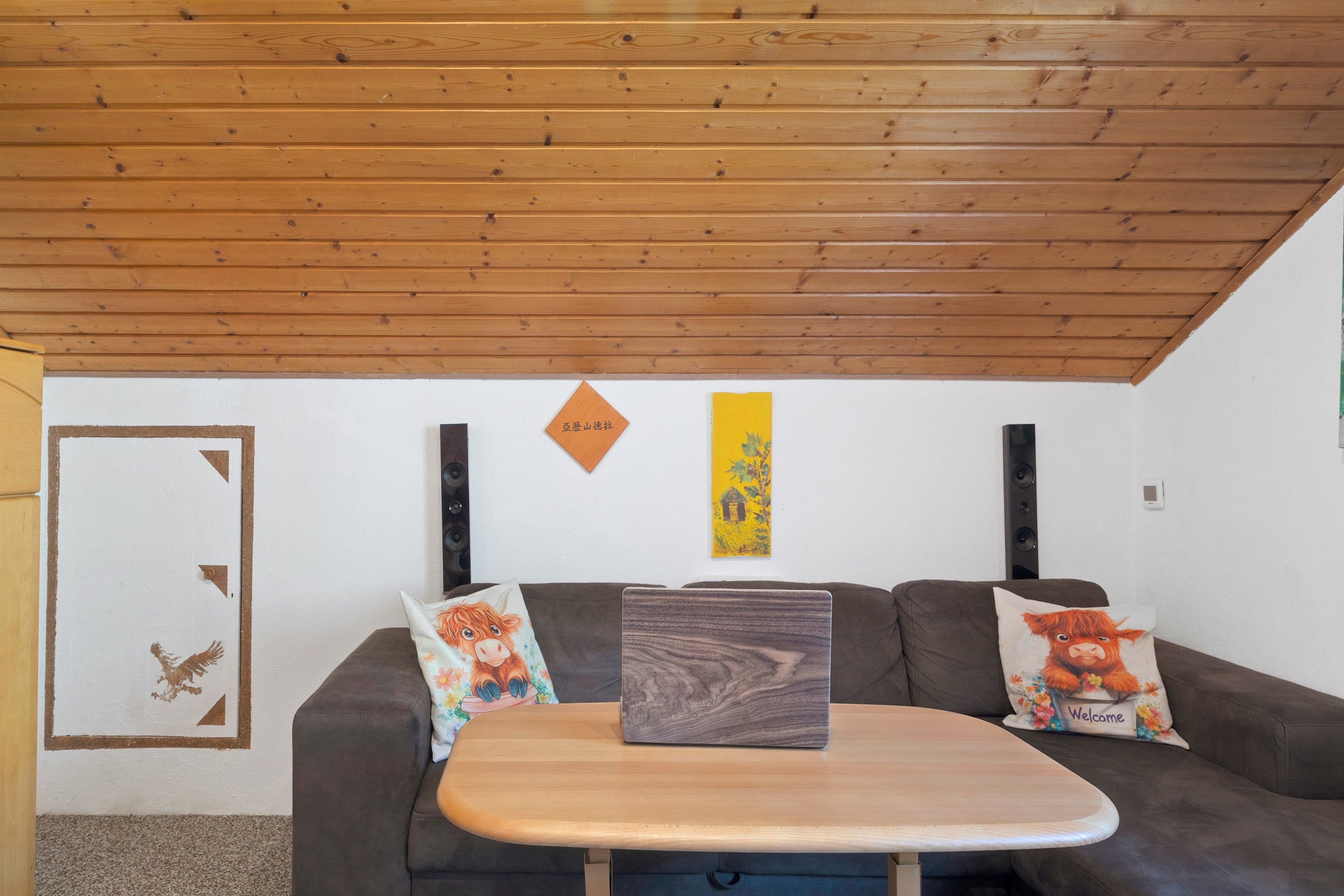 Une pièce avec un plafond en bois, un canapé agrémenté de deux coussins décoratifs, une petite table et un mur orné d’un tableau encadré et d’un panneau jaune vertical.
