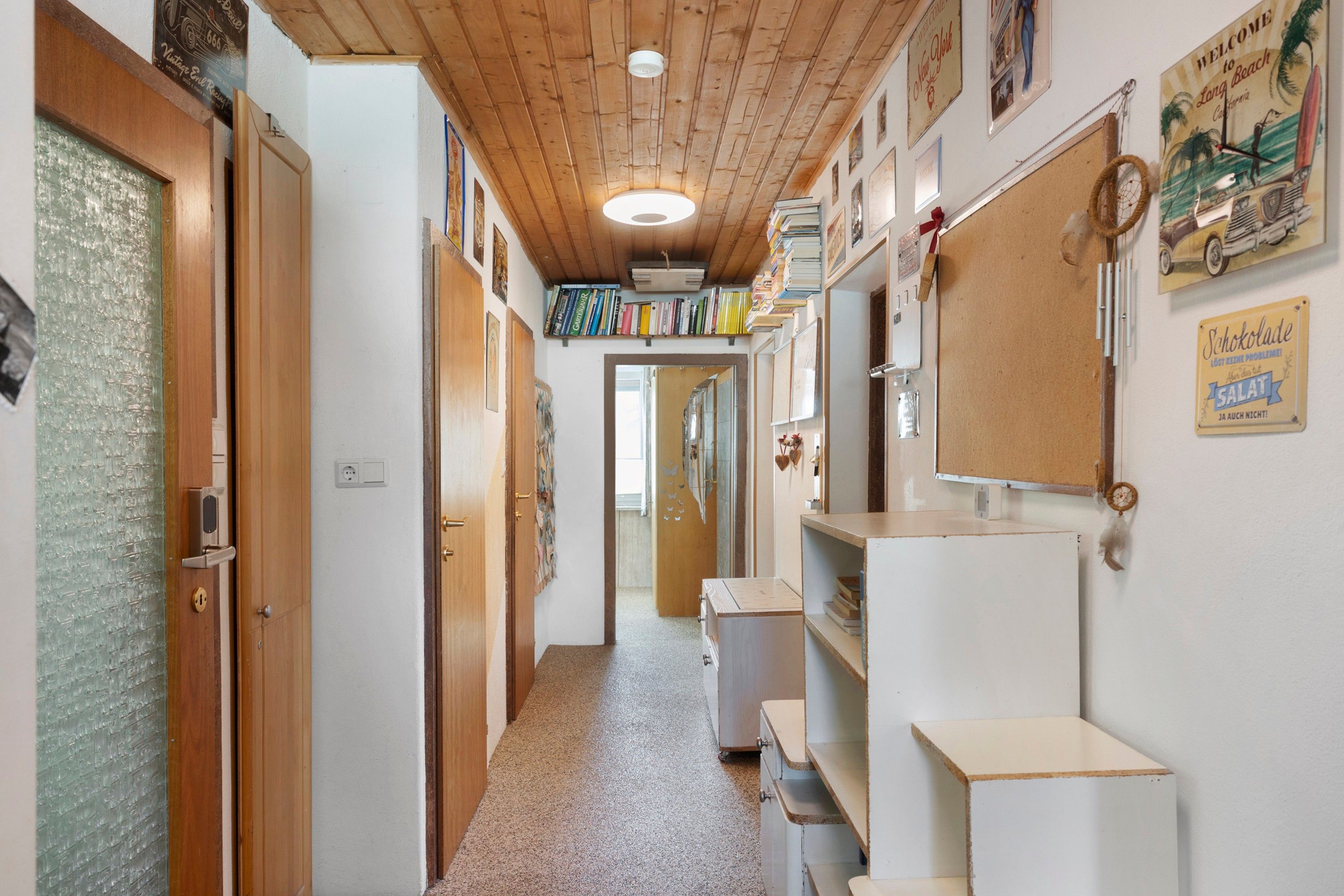 A narrow hallway with wooden ceiling beams, a door with a frosted glass panel, and a wall with various signs and decorations.