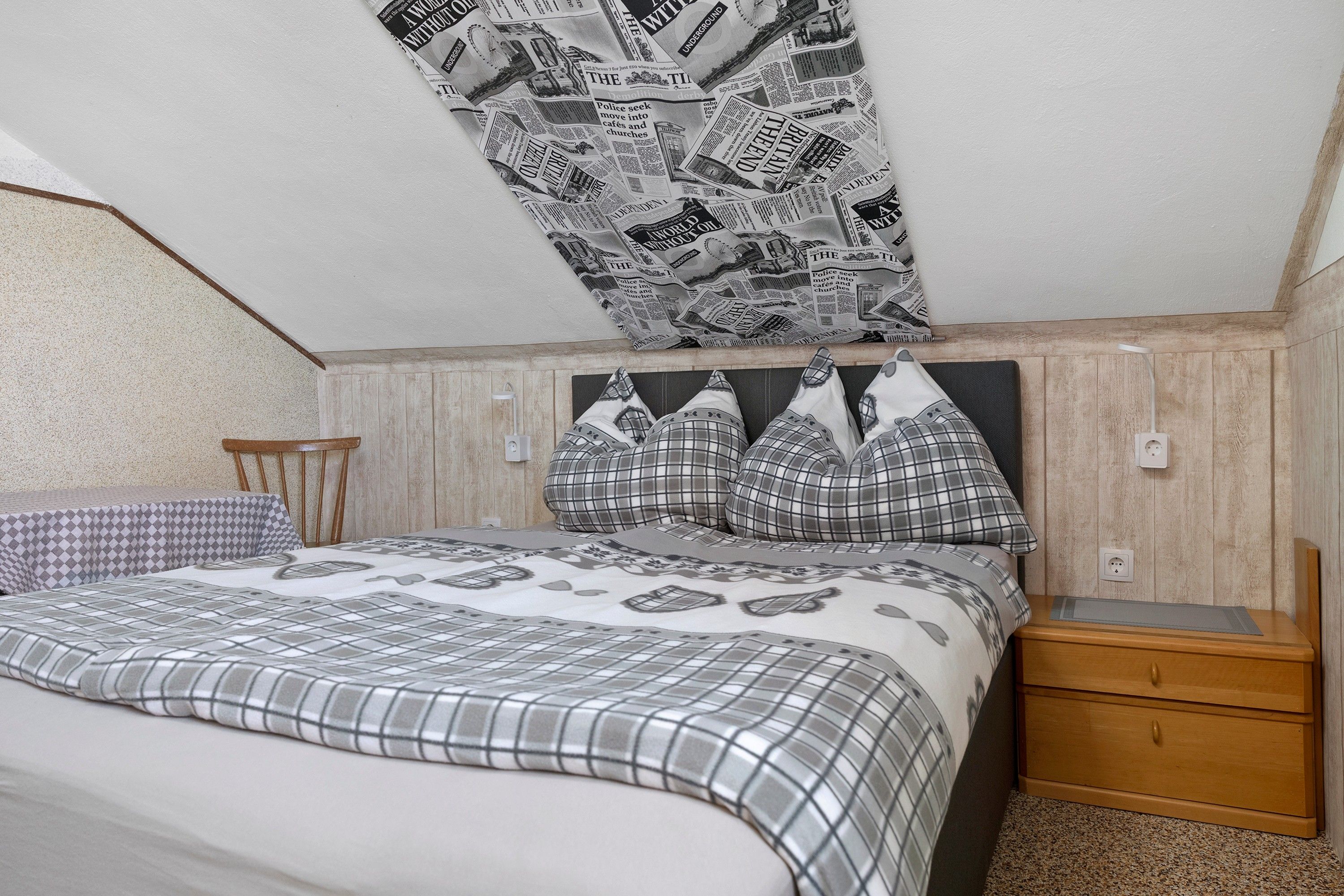 A bedroom with a sloped ceiling, a bed with a patterned bedspread, a nightstand, and a wall-mounted shelf.