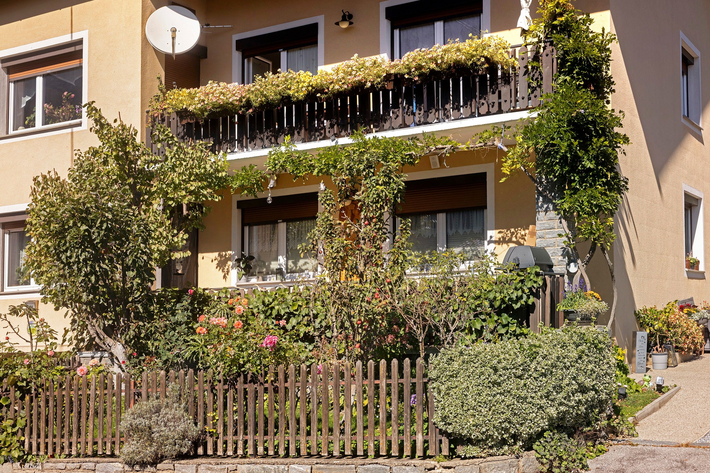 A multi-story building with balconies covered in flowering plants, a stone wall, and a wooden fence.
