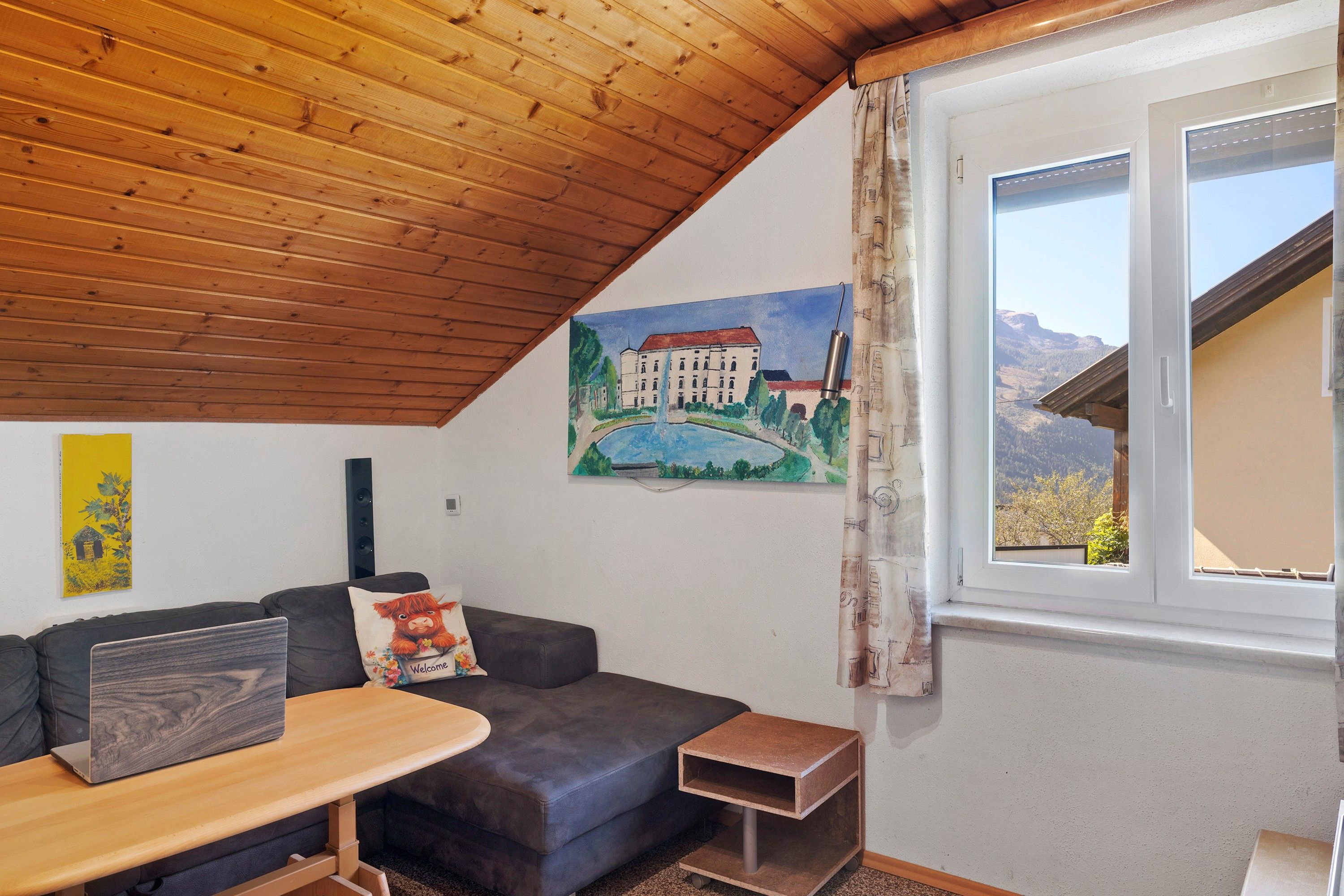 Una habitación acogedora con techo de madera, un sofá gris, una pequeña mesa con una computadora portátil, un cuadro de un edificio y una ventana con vistas a las montañas.