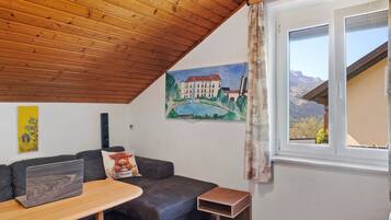 Una habitación acogedora con techo de madera, un sofá gris, una pequeña mesa con una computadora portátil, un cuadro de un edificio y una ventana con vistas a las montañas.
