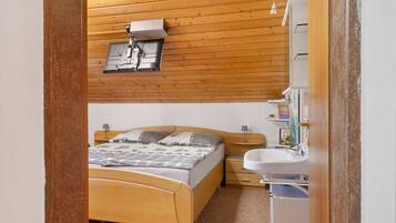 A bedroom with a wooden bed, a nightstand, a wall-mounted shelf, and a bathroom with a sink and mirror.