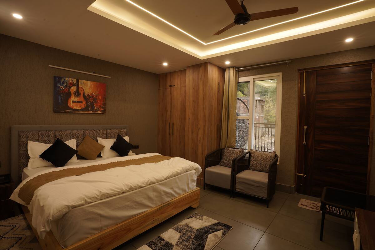 A bedroom with a bed, a ceiling fan, a window with curtains, a wooden door, and a painting on the wall.