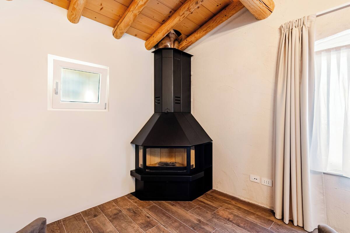 A corner room with a black wood-burning stove, a small window, and a wooden ceiling.