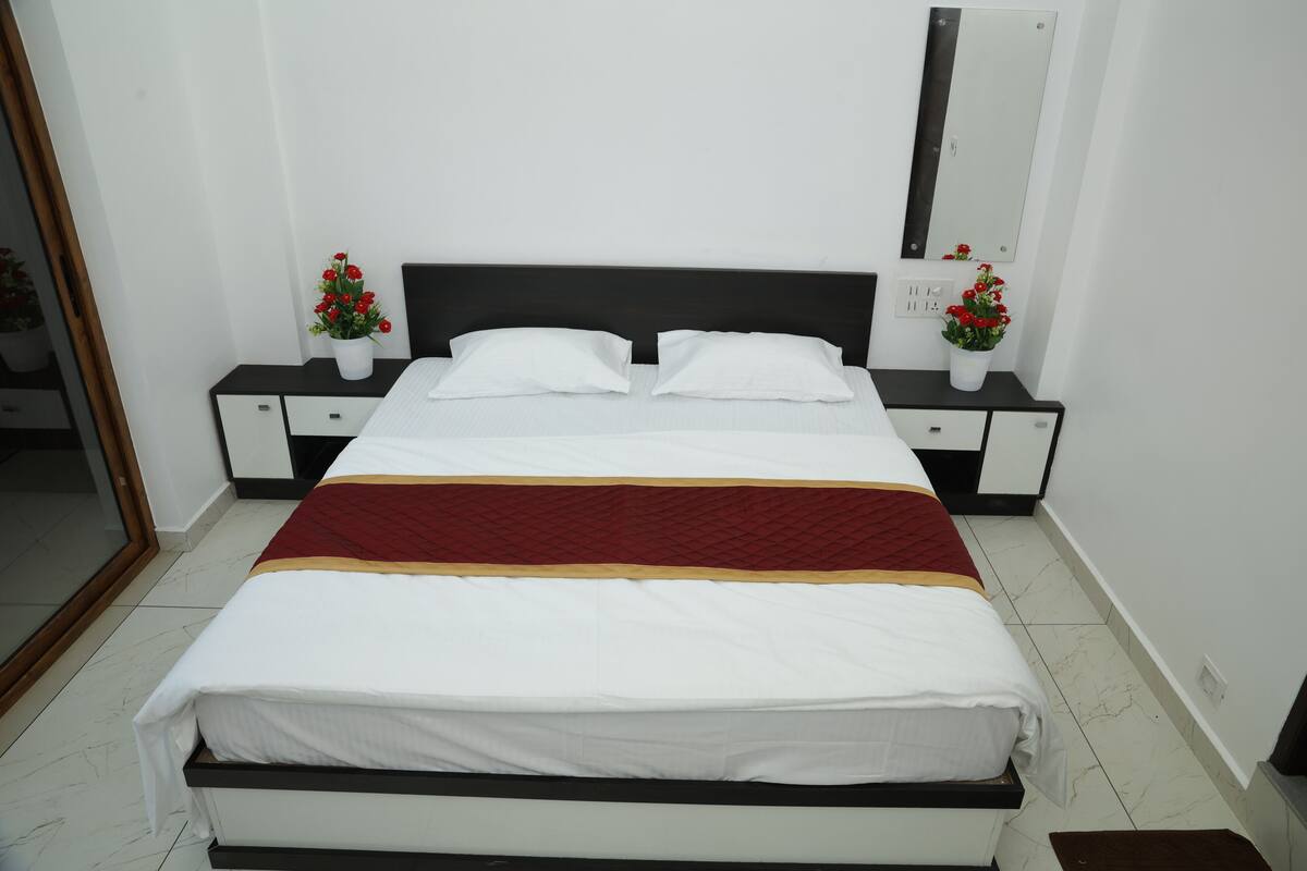 A neatly made bed with white and red bedding, two bedside tables with flower vases, a large mirror on the wall, and a tiled floor.