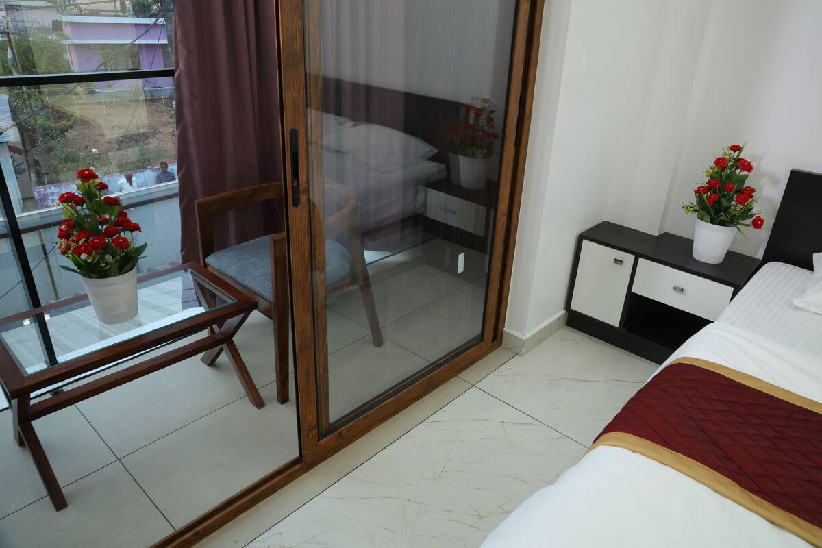A hotel room with a bed, a glass-enclosed sliding door, a small table with a potted plant, and a nightstand with a lamp.