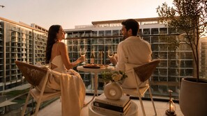 A couple seated at a table with drinks, a book, and a candle on a balcony with a city view. - Azizi Riviera Studio with Pool & Parking (Dubai)