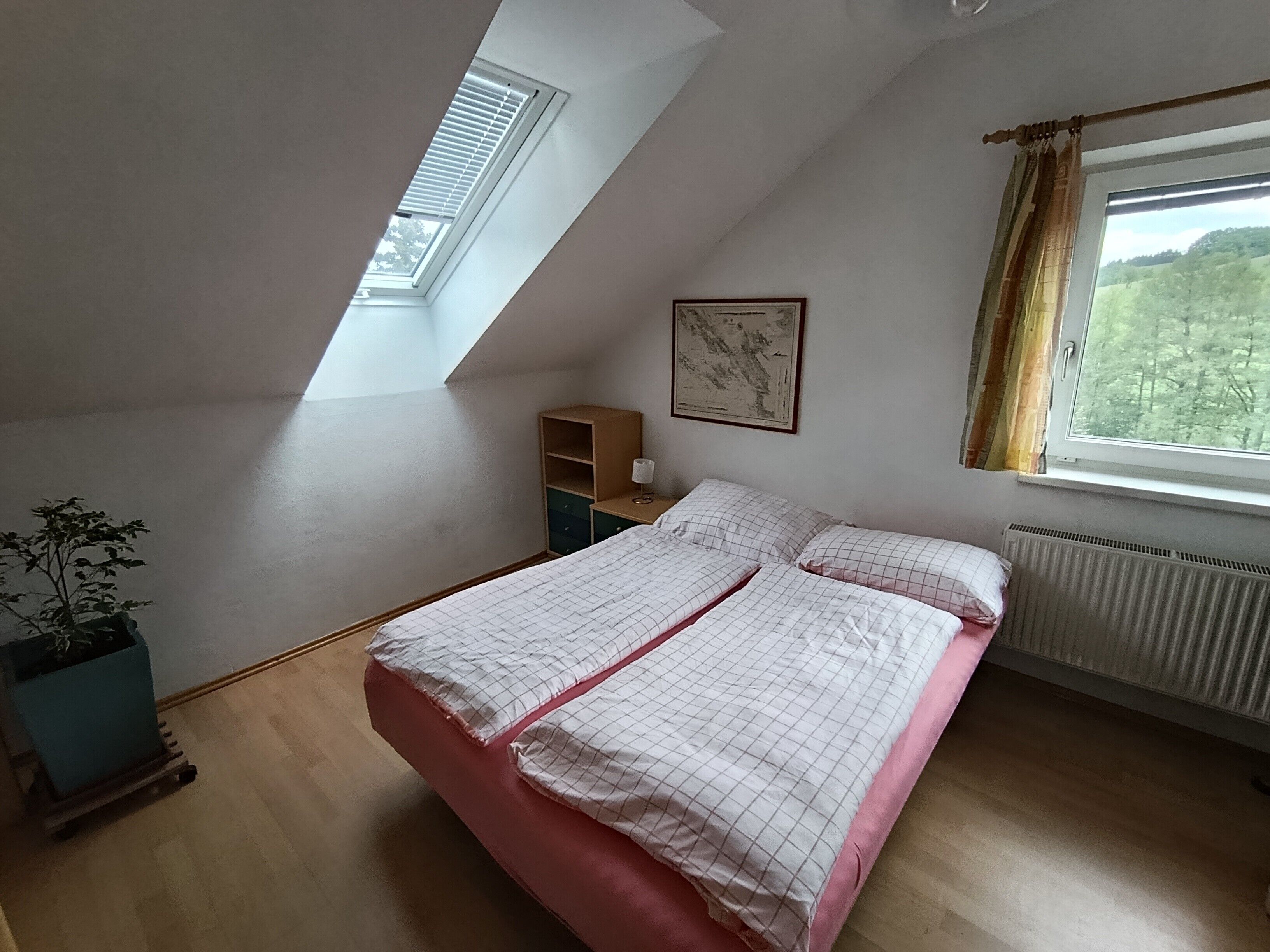 Une chambre avec un plafond en pente, un puits de lumière, un lit avec un matelas rose, une table de chevet, un radiateur et une fenêtre avec des rideaux.