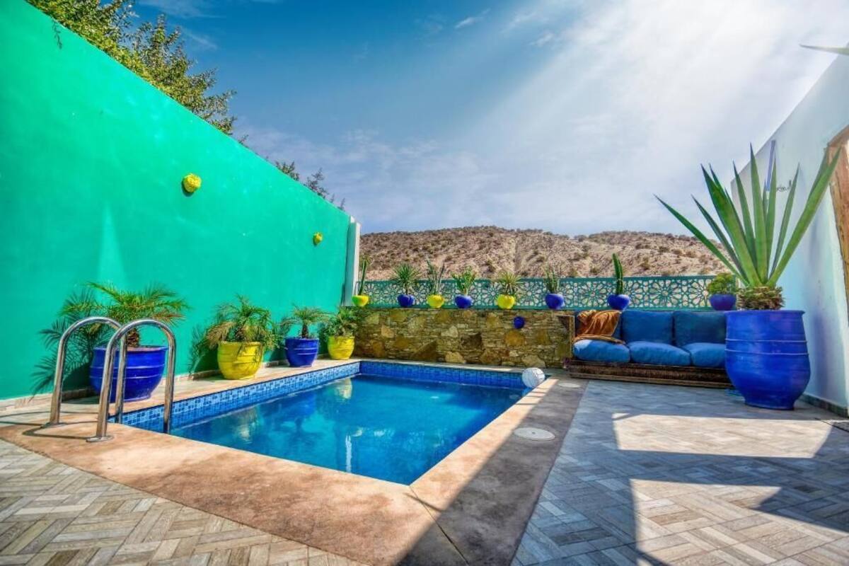 A swimming pool with a stone wall and potted plants, a blue sofa, and a view of hills.