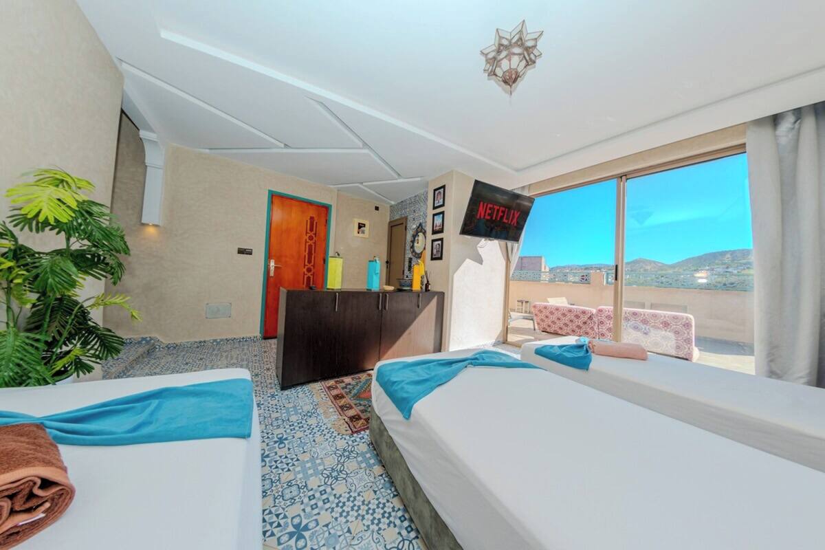 A hotel room with two beds, a TV, a desk, a plant, and a view of the landscape.