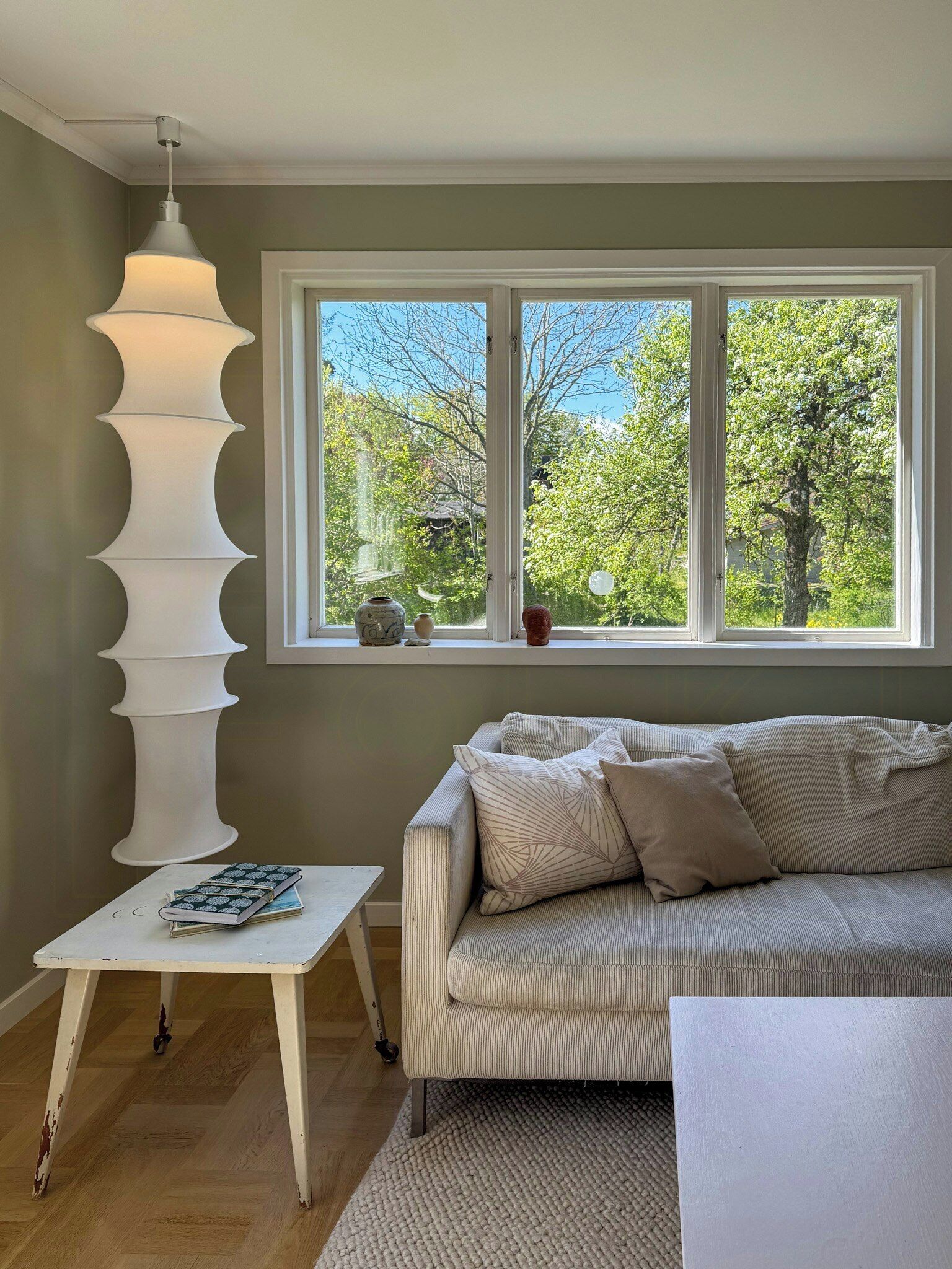 Ett rum med ett stort fönster, ett vitt bord med ett anteckningsblock, en beige soffa och en hög, vit lampa.
