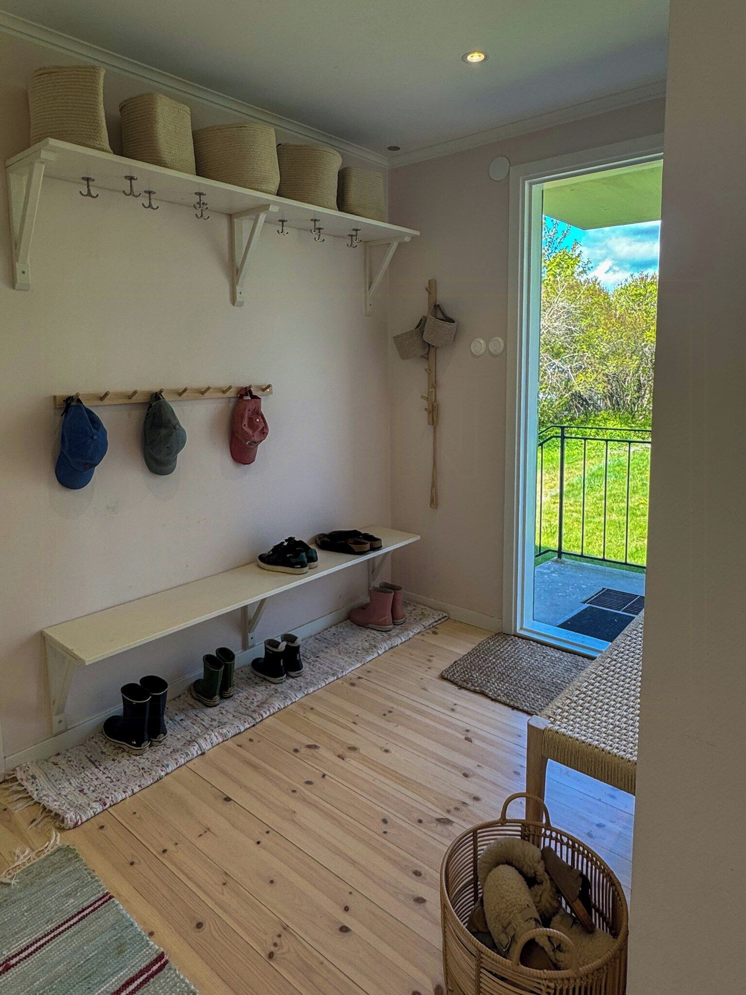 A room with a wooden floor, a bench with shoes, a basket with items, and a view of greenery outside.