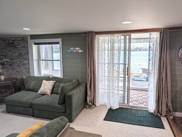A living room with a green sofa, a window with a view of a body of water, and a stone wall. - NEW!  Villa Adonis - Lakefront, Dog-Friendly, and Gorgeous!  Includes NEW 6-PERSON HOT TUB, Central Air, and Water Toys! (National City)