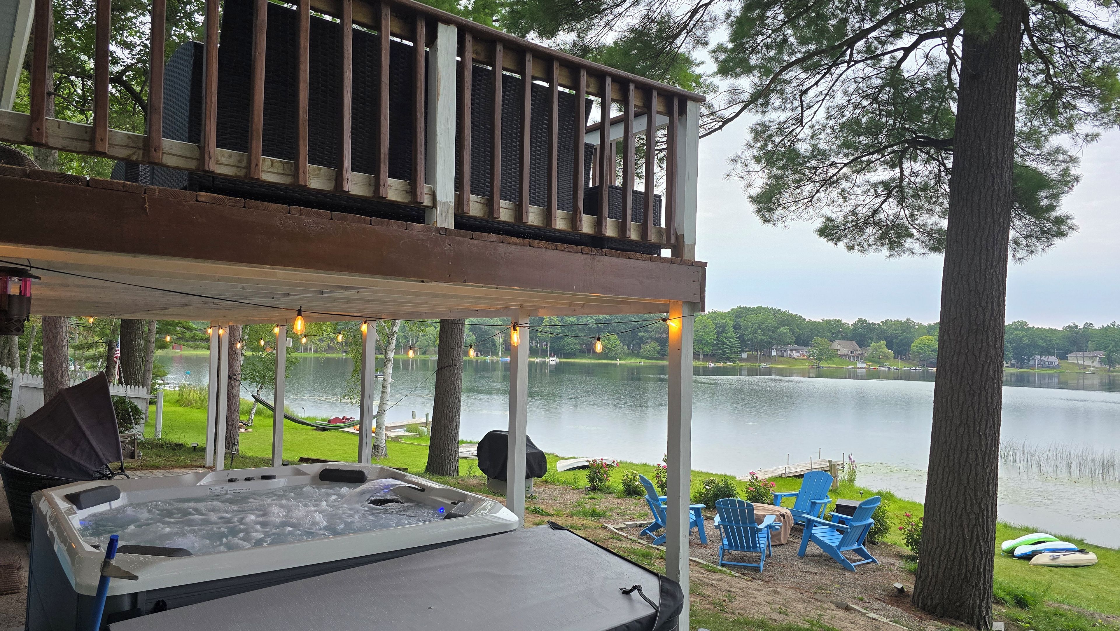 A hot tub with a view of a lake, outdoor chairs, and a deck with string lights.