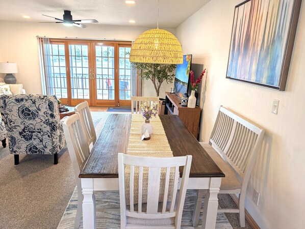 A dining area with a wooden table, white chairs, a woven pendant light, and a large window with a view.