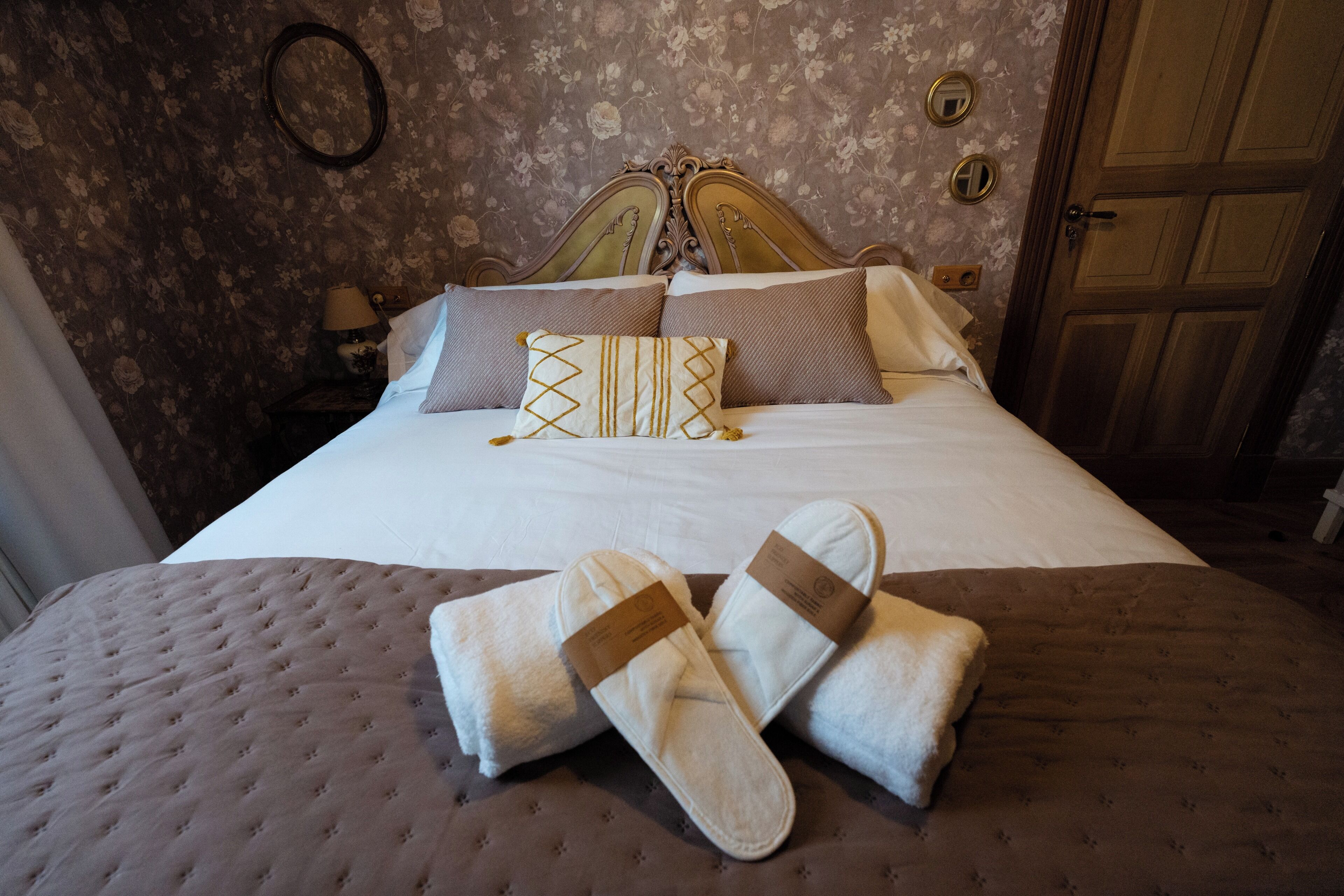 Una cama bien hecha con almohadas, cabecera y pantuflas sobre una colcha con textura.