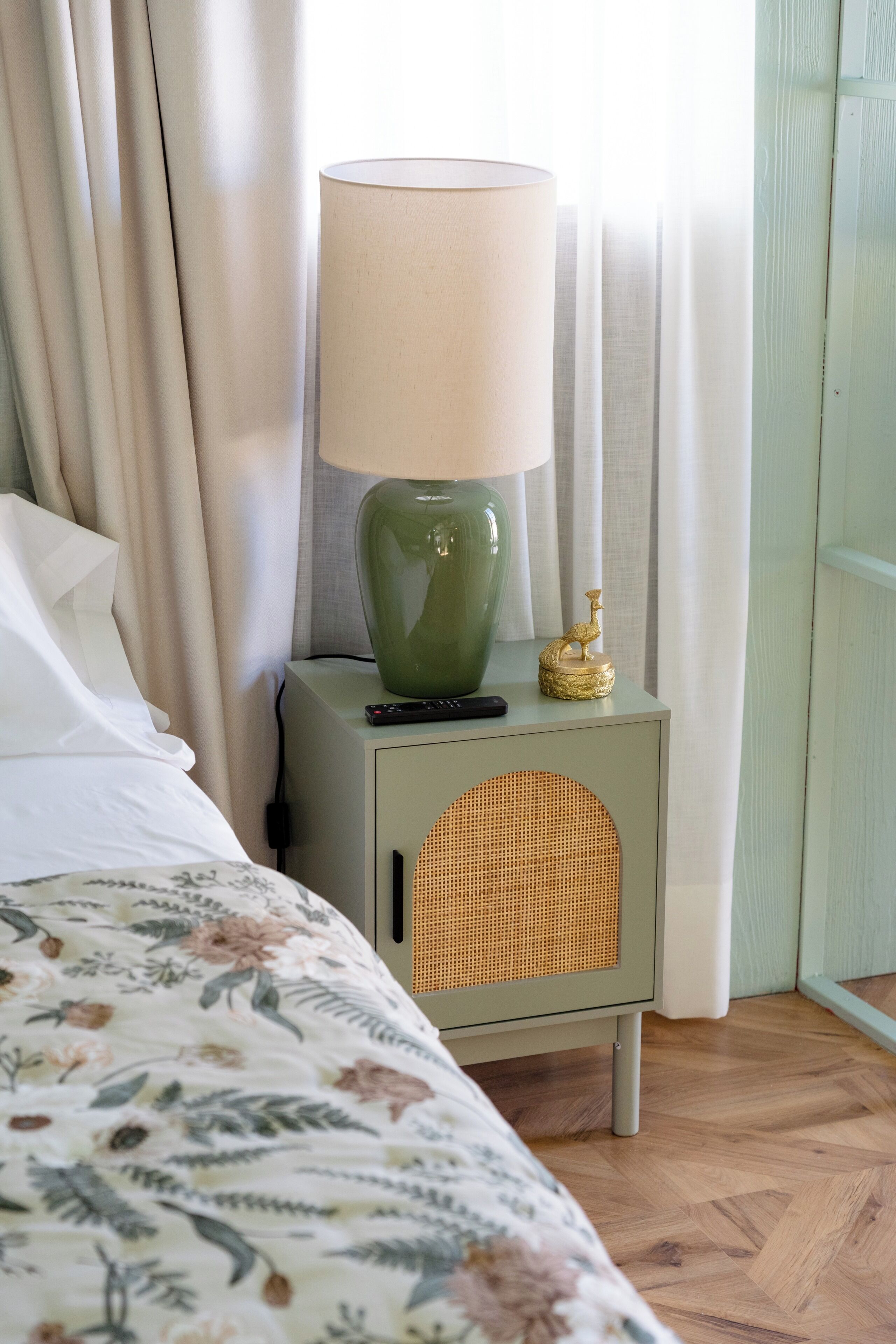 A neatly made bed with a floral bedspread, a green bedside lamp with a beige shade, and a wicker speaker on a light green nightstand.