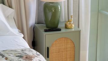 A neatly made bed with a floral bedspread, a green bedside lamp with a beige shade, and a wicker speaker on a light green nightstand.