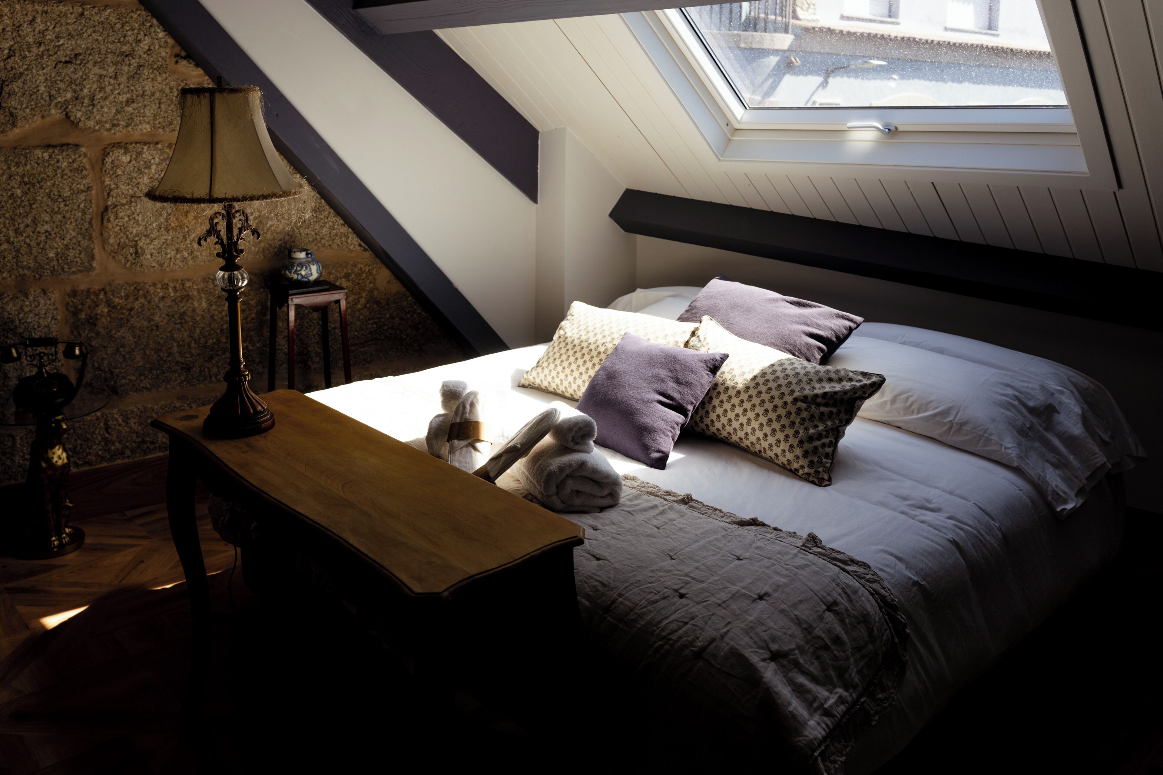 Een slaapkamer met een schuine kap, een houten nachtkastje, een lamp, een bed met opvallende kussens en een stenen muur.
