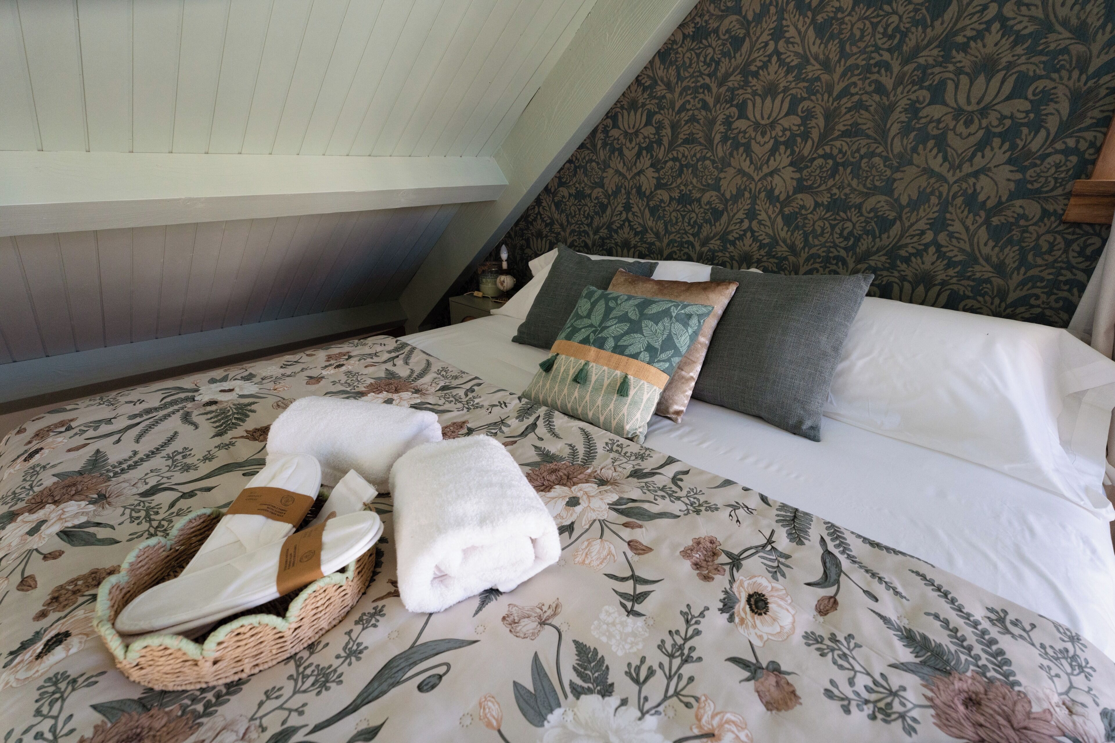 A bedroom with a floral bedspread, a basket of folded towels, and a nightstand with a lamp.