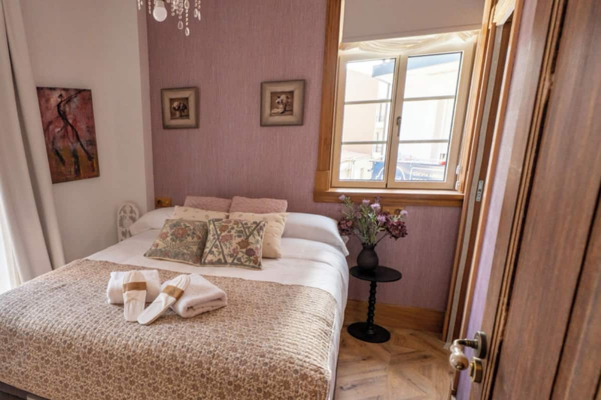 A neatly made bed with pillows and a blanket, a small table with a vase, and a window with curtains.