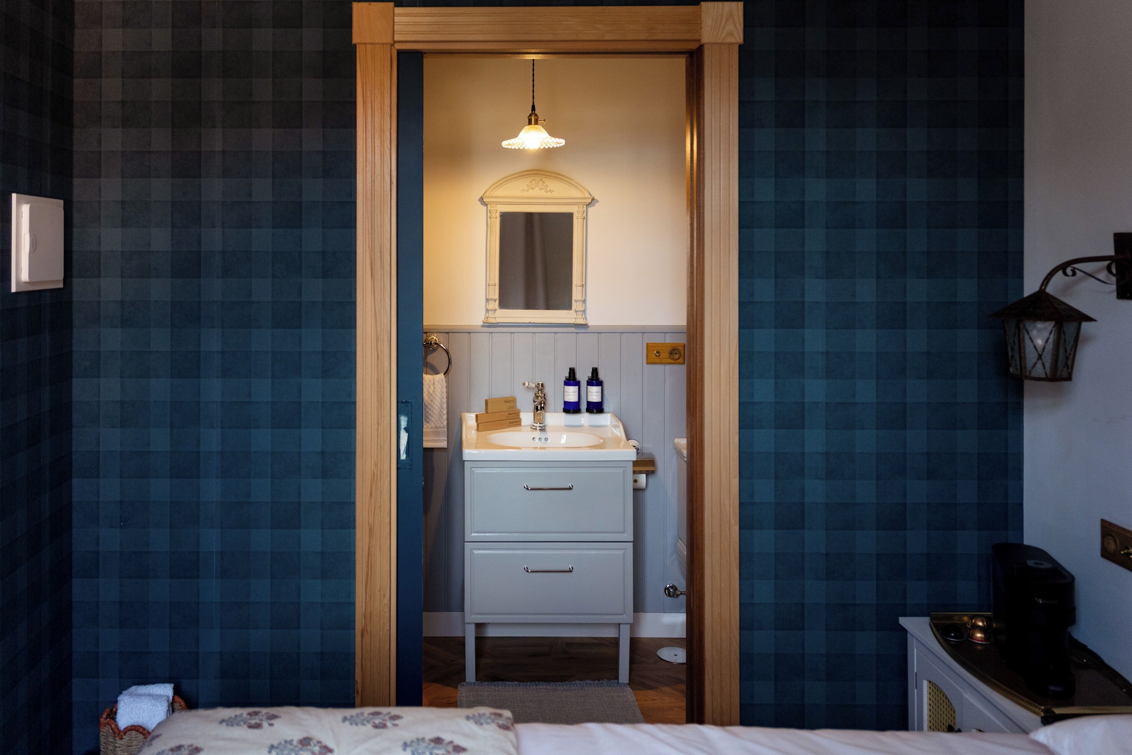 Une chambre d’hôtel avec un lit, une porte en bois donnant sur une salle de bain équipée d’un lavabo et d’un miroir, et une petite table avec une lampe.
