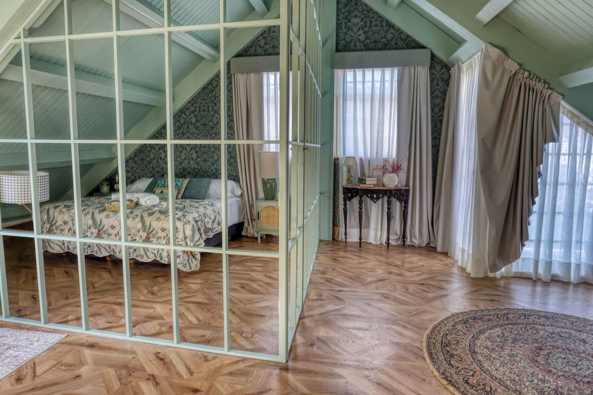 A room with a sloped ceiling, a bed with a floral bedspread, a small table with a lamp, and a large window with curtains.