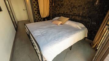 Un letto con struttura metallica, biancheria bianca e un cuscino color beige in una stanza con parete decorata e una porta sullo sfondo.
