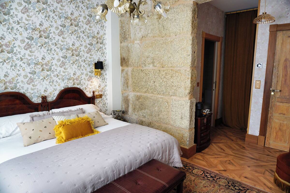 A bedroom with a bed, a wooden bench, a stone wall, a wooden door, and a patterned rug.
