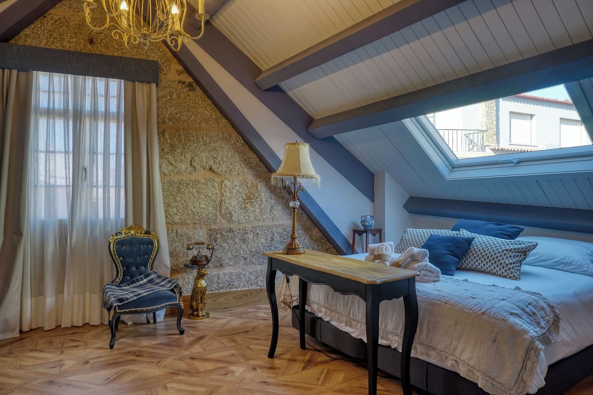 A bedroom with a sloped ceiling, a bed with pillows, a chair, a table, and a lamp.