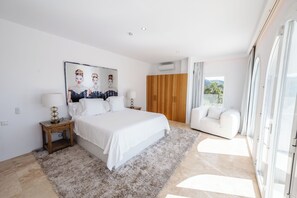 A bedroom with a large bed, a wooden wardrobe, a white sofa, and a view through a glass door. - WHITE OASIS Ibiza Villa (Sant Llorenc de Balafia)