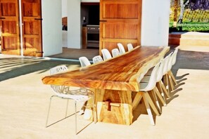 A long wooden dining table with chairs, set outdoors with a view of a building and greenery. - WHITE OASIS Ibiza Villa (Sant Llorenc de Balafia)