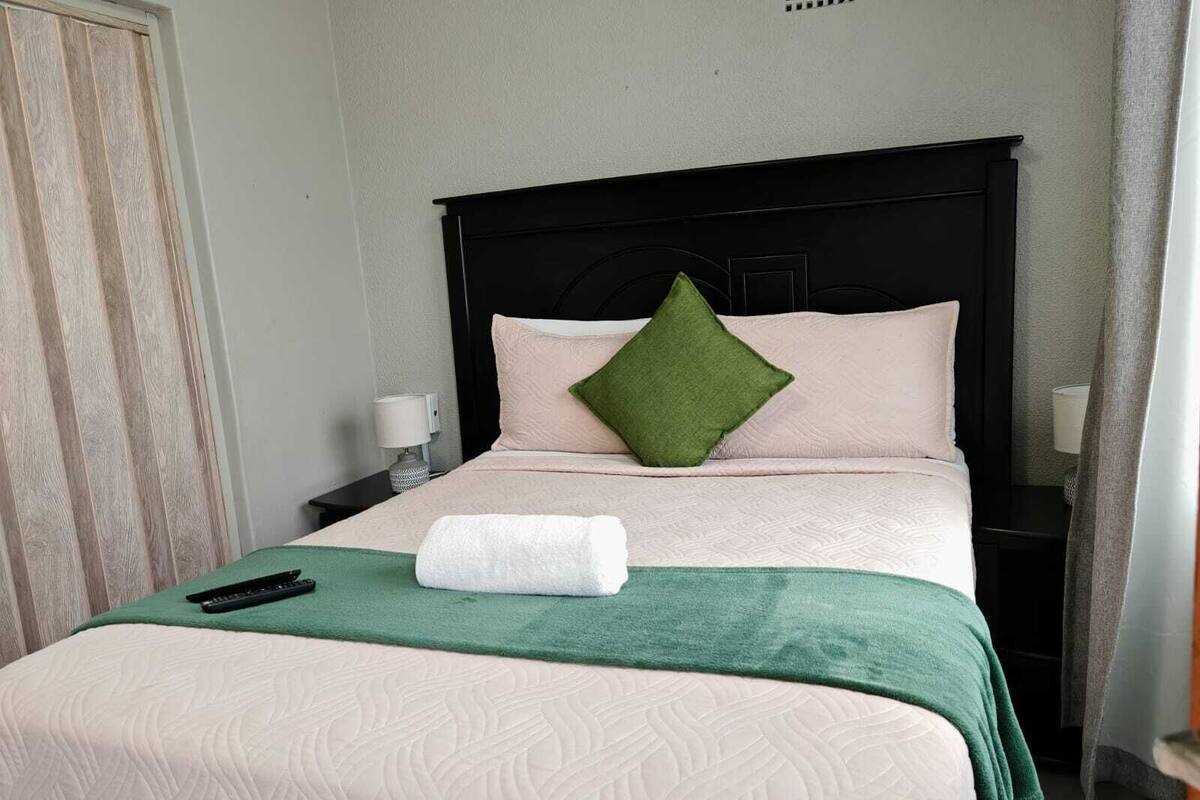 A neatly made bed with a green throw, a remote control, and a rolled towel on a white bedspread.