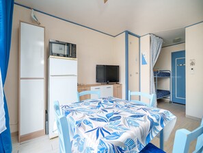 Ein kleines Zimmer mit Esstisch, Stühlen, Fernseher, Kühlschrank und einem Etagenbett.