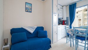Une pièce avec un canapé bleu, un petit coin cuisine et une vue sur les immeubles par la fenêtre.