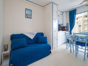 Ein Zimmer mit blauem Sofa, einer kleinen Küchenzeile und Blick auf Gebäude durch ein Fenster.