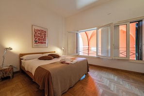 Ein Schlafzimmer mit Bett, Nachttisch, Lampe, Fenster mit Blick auf Orange und einem Wandbild.