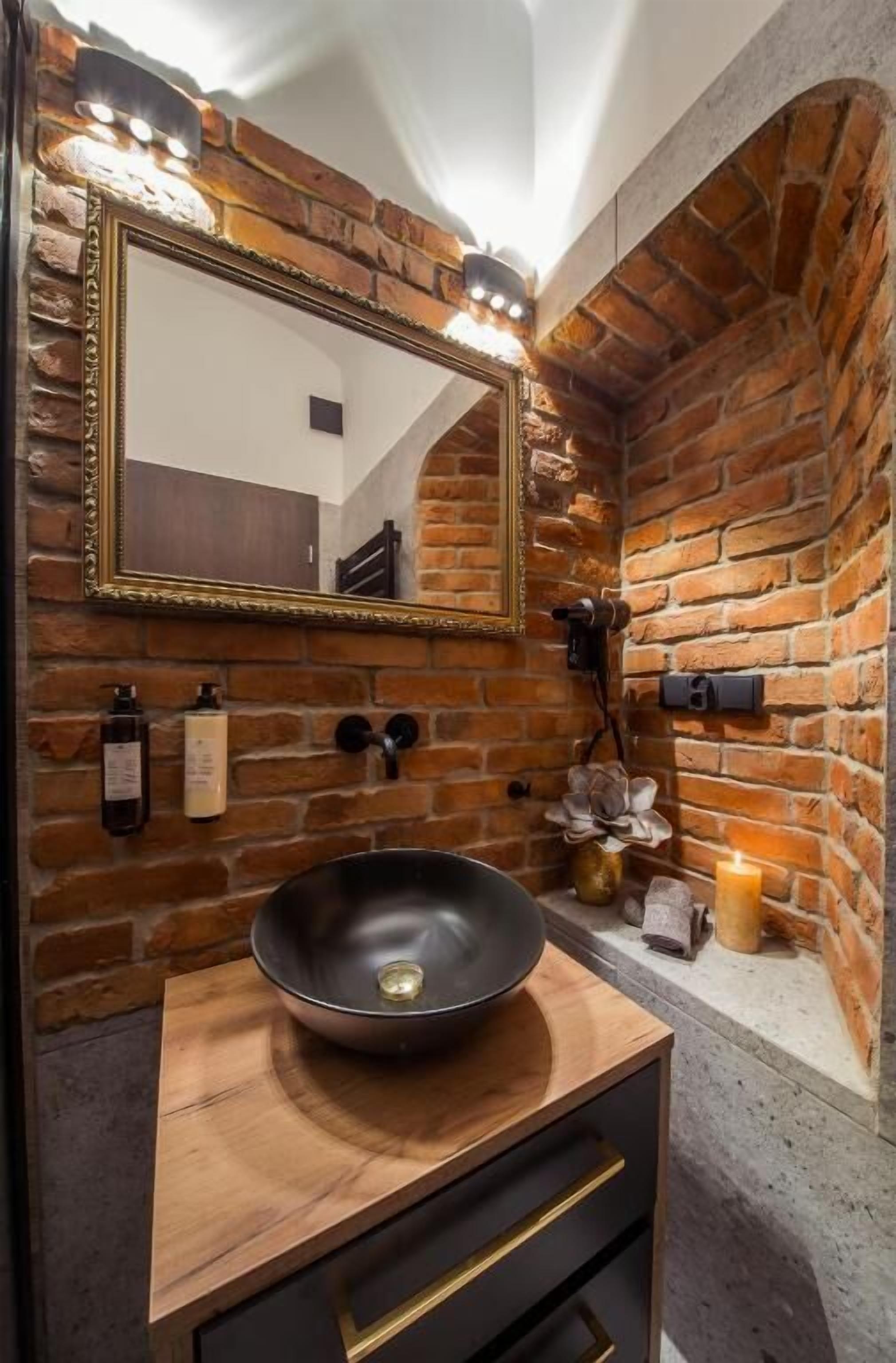 Un baño con tocador de madera, un lavabo negro, un espejo con marco dorado y una pared de ladrillo.