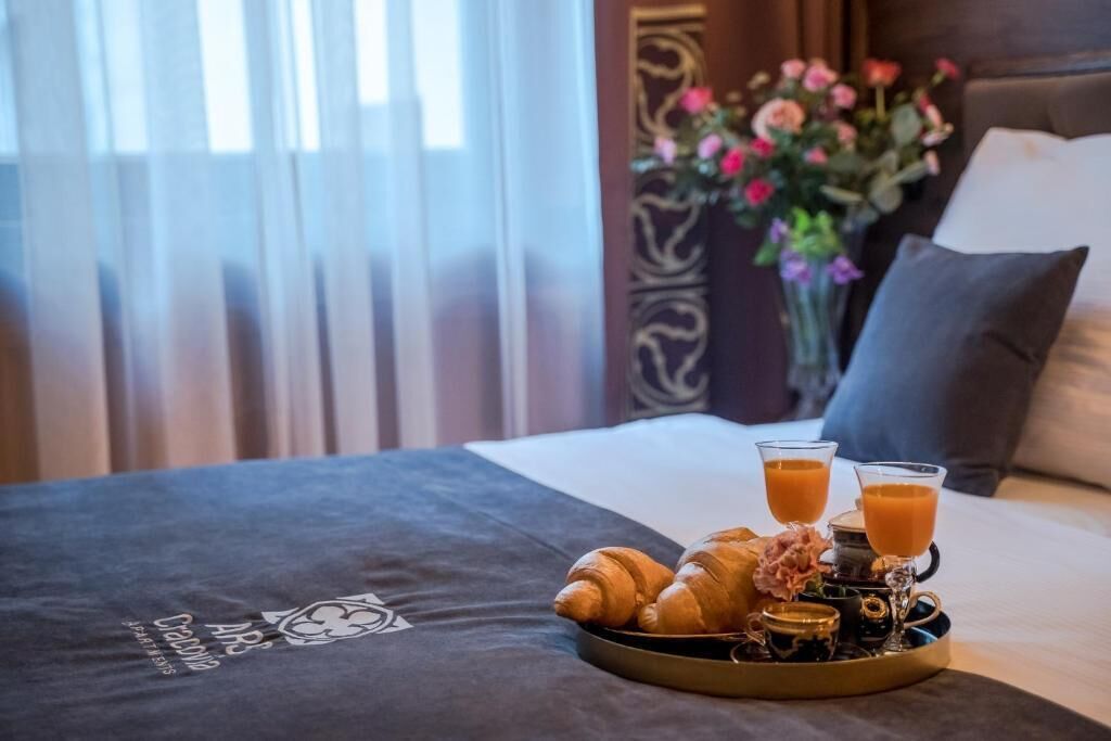 Una cama con una bandeja de cruasanes y jugo de naranja, un jarrón de flores y una colcha oscura con un logotipo.