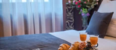 Ein Bett mit Tablett mit Croissants und Orangensaft, einer Blumenvase und einer dunklen Bettdecke mit einem Logo.